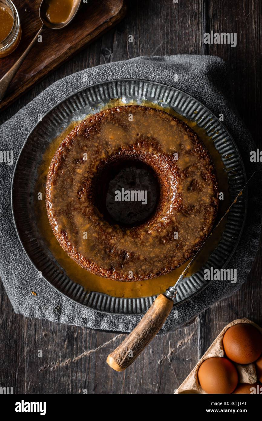 Budino di spugna di sciroppo dorato appiccicoso in una sagoma ad anello con glassa al caramello su un tavolo di legno scuro per un dolce accogliente. Foto Stock
