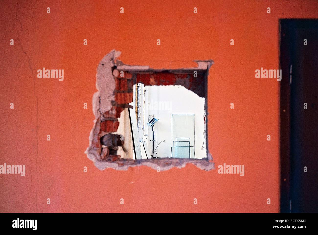 Foro quadrato in una parete arancione che rivela il vetro della finestra appoggiato contro la parete interna, catturato in pellicola analogica che mette in risalto texture e luce. Foto Stock