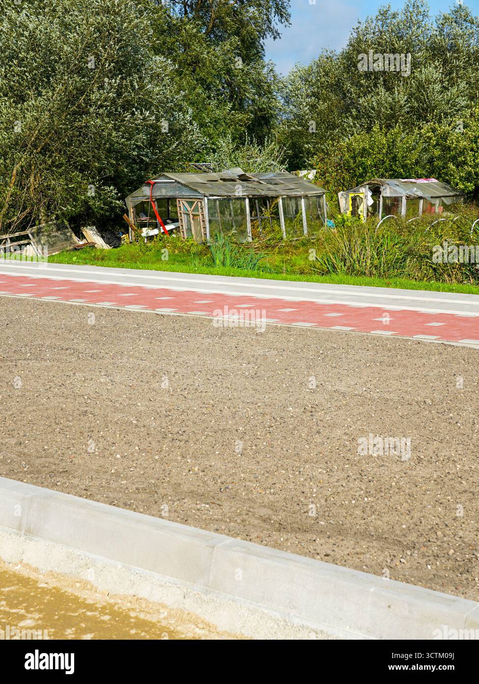 Una moderna passerella pavimentata di rosso contrasta con vecchie serre abbandonate circondate da vegetazione selvaggia, simboleggiando il cambiamento tra vecchia e nuova infrastruttura Foto Stock