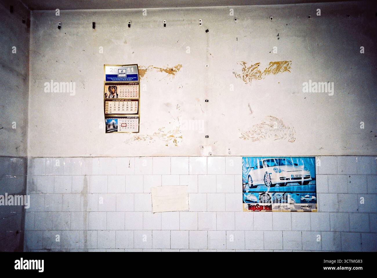 Parete piastrellata bianca in una fabbrica abbandonata adornata da un vecchio calendario e una foto di auto sportive, catturata in un flash analogico. Foto Stock