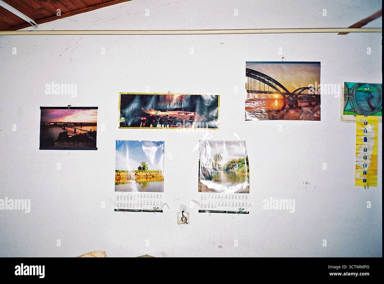 Parete bianca in una fabbrica abbandonata adornata da vecchi calendari e fotografie di ponti, catturata con flash analogico per texture e atmosfera. Foto Stock