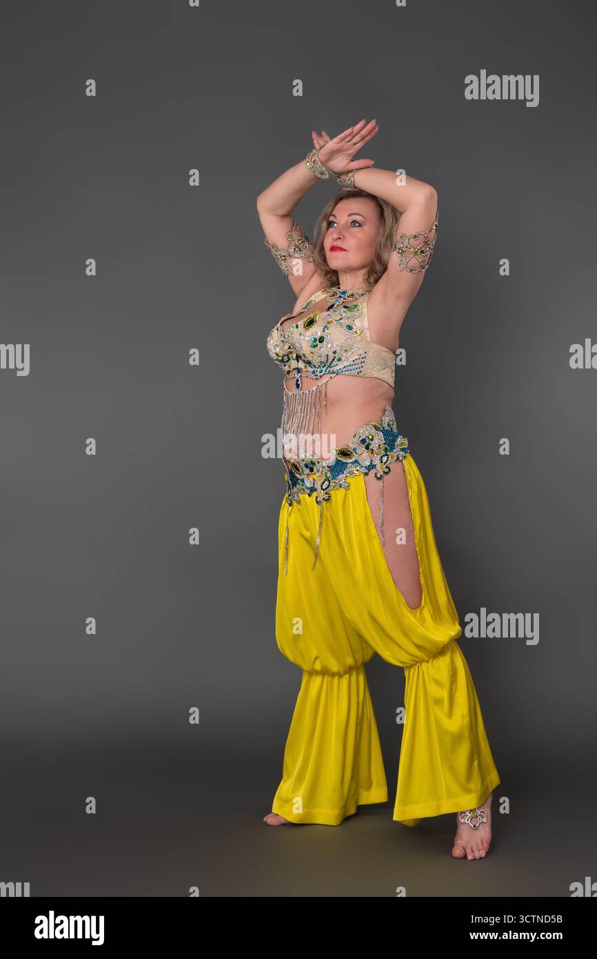 Donna curvy danzatrice della generazione X che indossa il costume da ballo: I pantaloni harem superiori e gialli si muovono a piedi nudi su fondo grigio Foto Stock