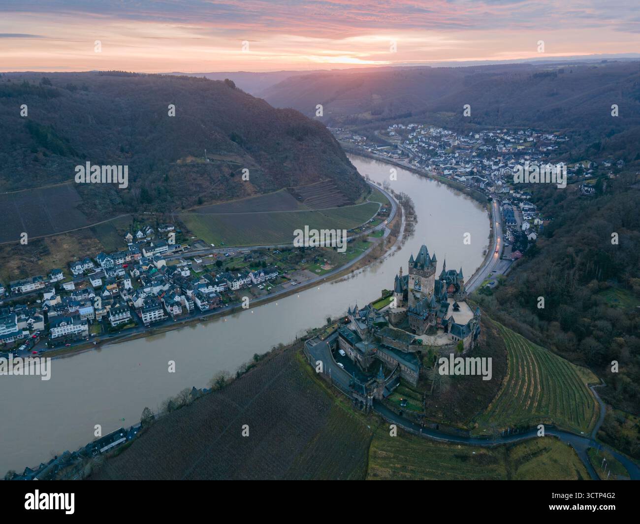 Vista aerea del castello di Cochem arroccato sopra il fiume Mosella mentre il sole sorge dietro nuvole rosa, illuminando il fiume e i vigneti circostanti Foto Stock