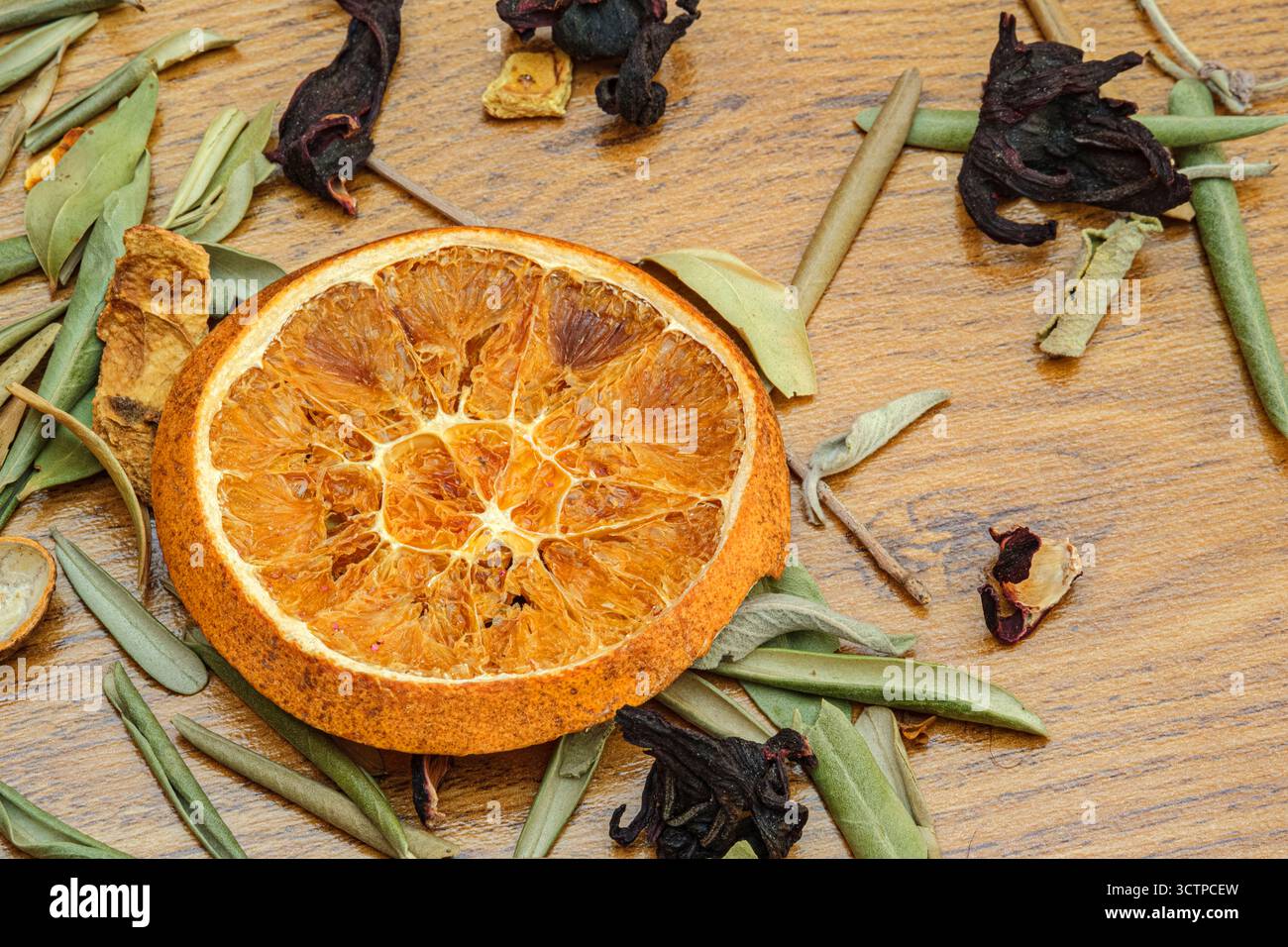Primo piano di una fetta di arancia secca circondata da erbe e foglie su una superficie di legno. La disposizione naturale riflette gli ingredienti del tè alle erbe, aromathe Foto Stock