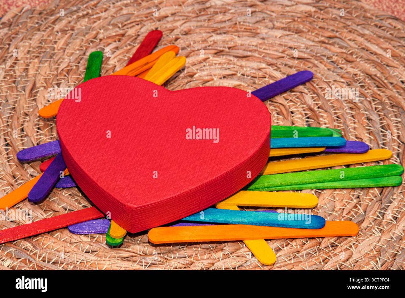Scatola rossa a forma di cuore posta sopra colorati bastoncini di legno in tonalità arcobaleno. La disposizione crea una scena giocosa e allegra. Foto Stock
