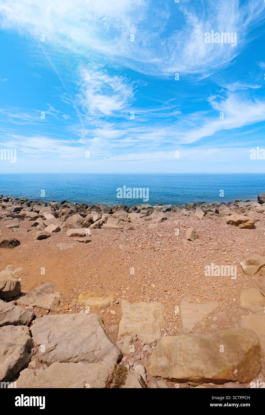Vista panoramica della costa rocciosa sotto un cielo azzurro luminoso con nuvole mostruose. Le pietre e i ciottoli incontrano l'orizzonte calmo del mare. Creare una pace. Foto Stock