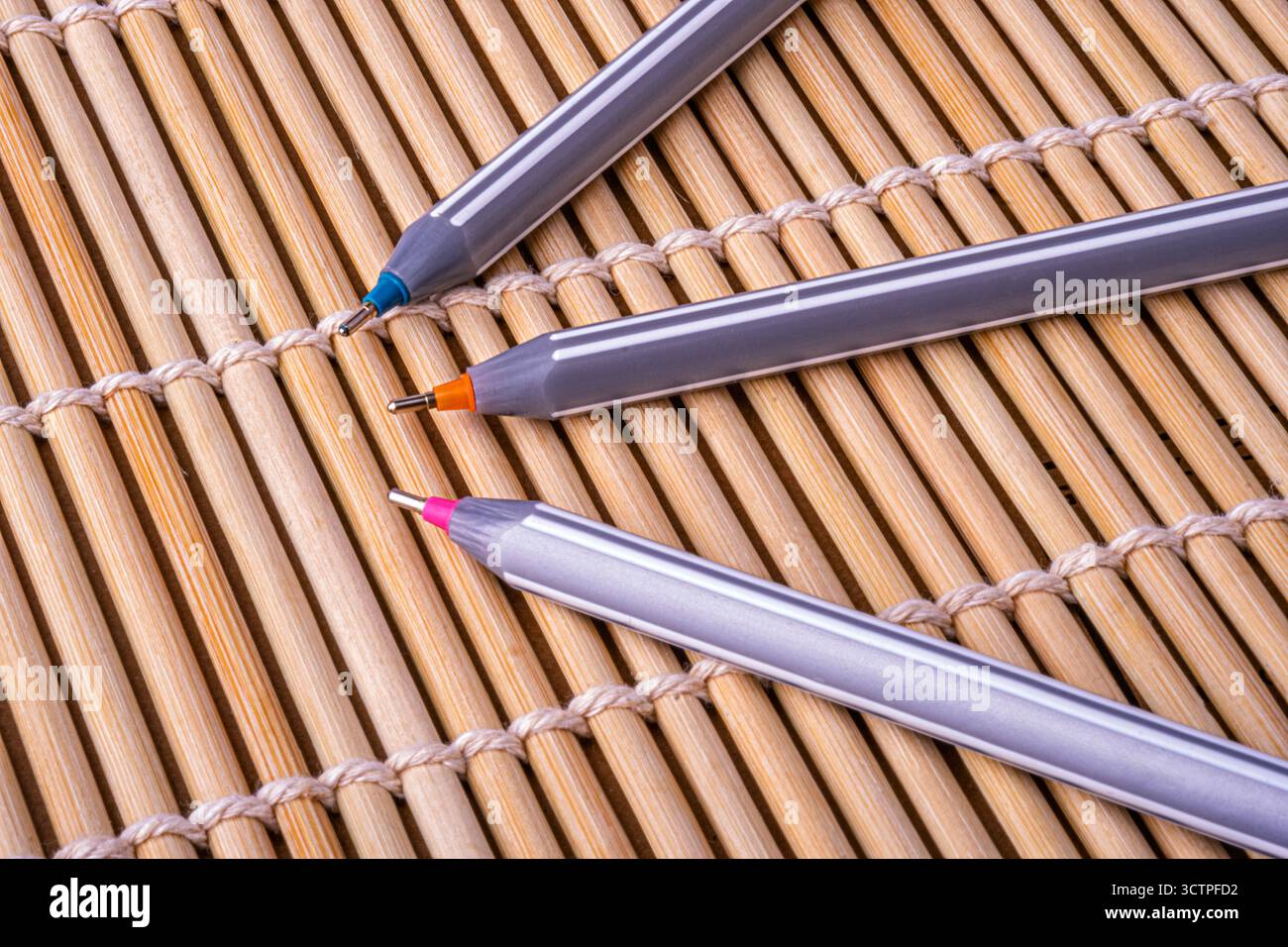 Tre penne a sfera con punte colorate sono posizionate diagonalmente sul tappetino in bambù. La struttura in legno naturale contrasta con il moderno design della cancelleria. Foto Stock