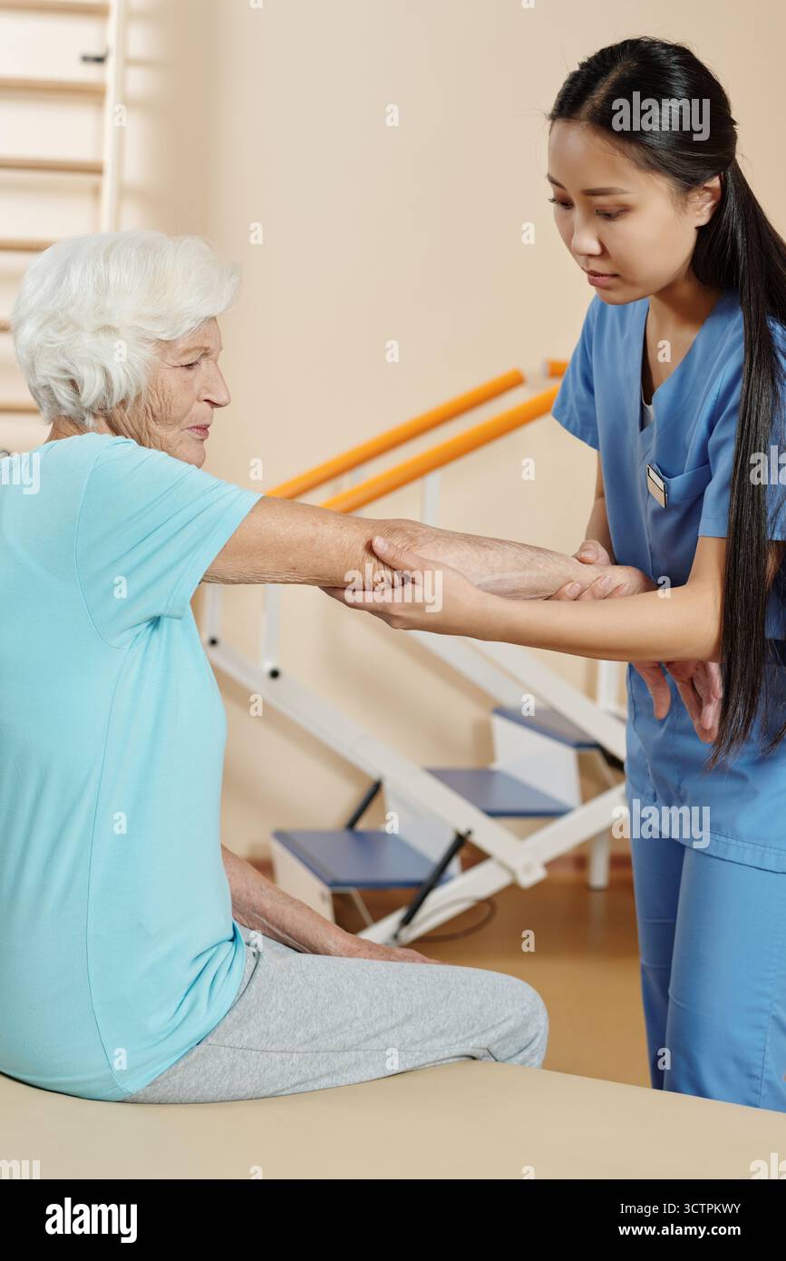 Giovane terapeuta fisica asiatica che fa il check-up di donna caucasica anziana seduta sul divano in ospedale di riabilitazione Foto Stock