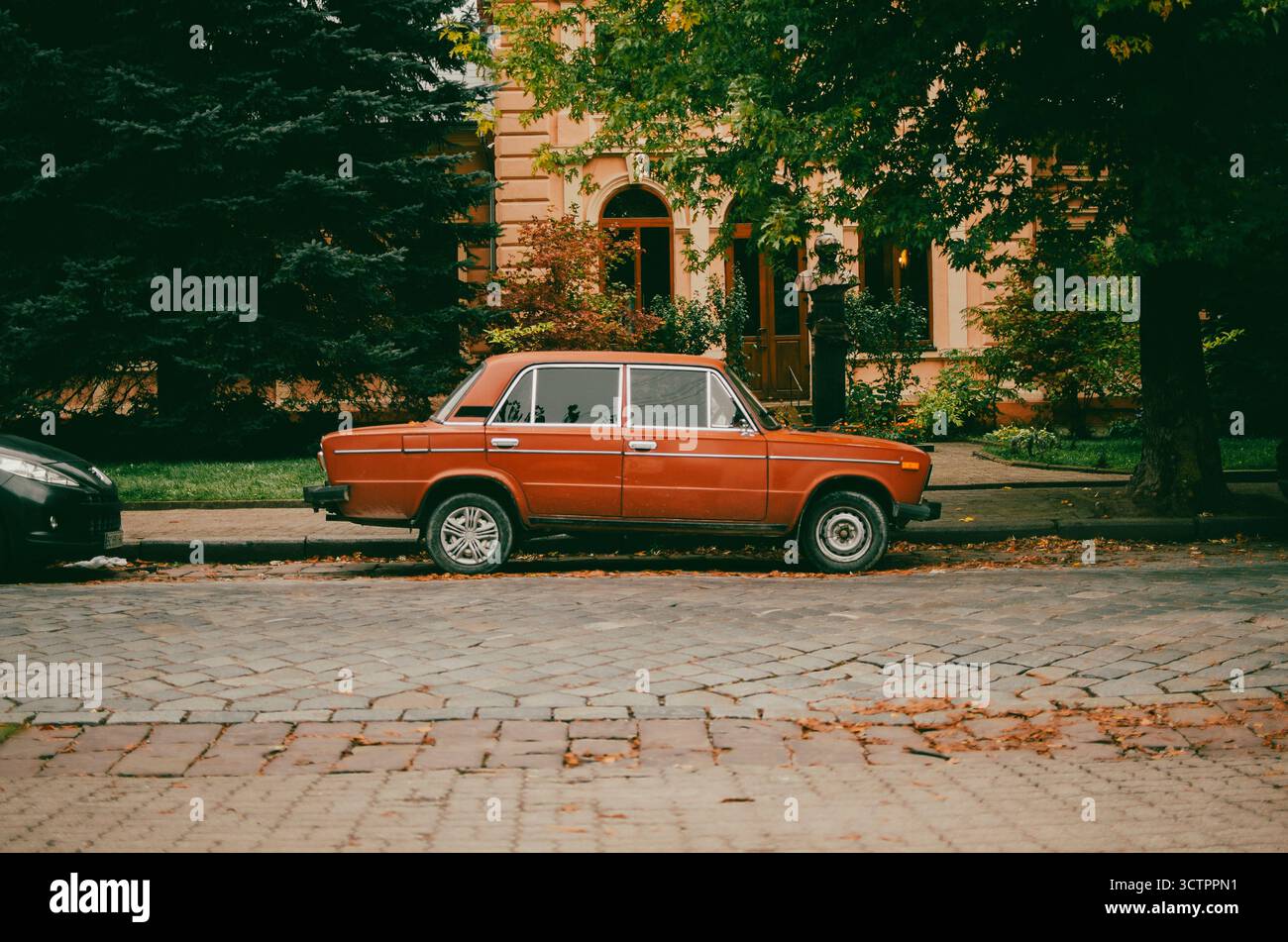 Berlina rossa d'epoca parcheggiata su una strada acciottolata di fronte a un edificio classico con finestre ad arco e lussureggianti alberi verdi. Atmosfera serena con Foto Stock