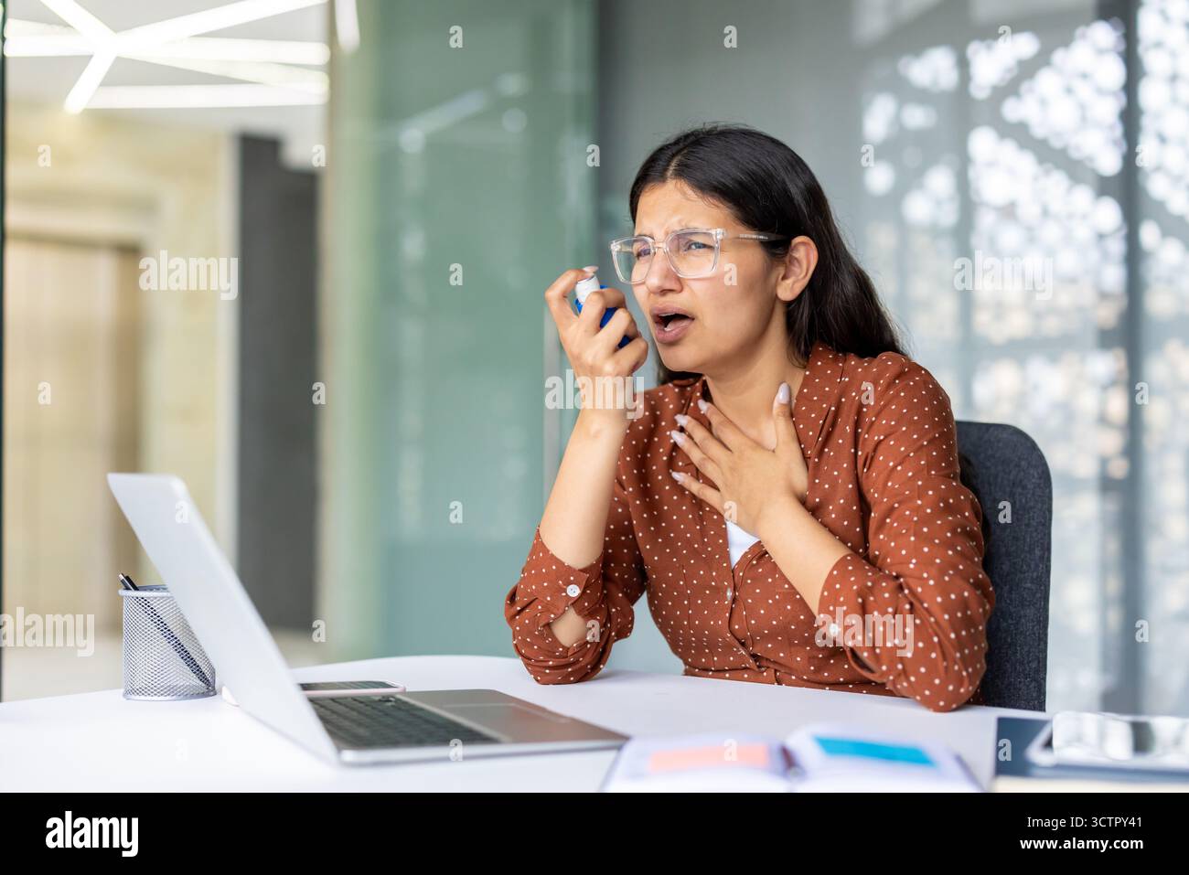 Giovane donna con un improvviso attacco di asma e difficoltà respiratorie, utilizzando un inalatore per alleviare i sintomi mentre lavora su un portatile in un ambiente di ufficio moderno Foto Stock
