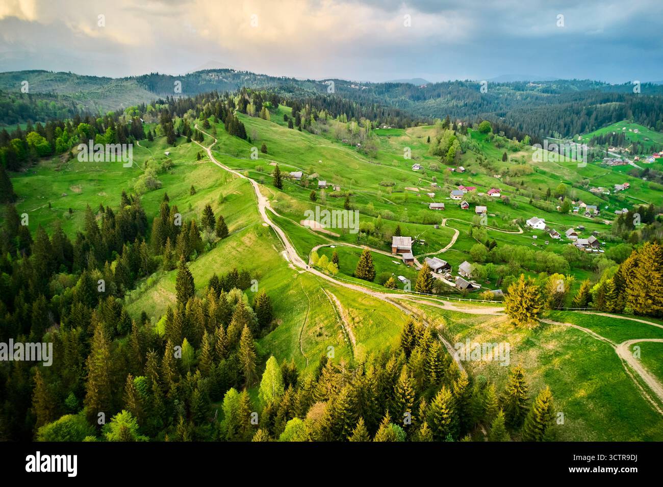 Fotografia aerea di un tranquillo paesaggio rurale in Ucraina, con colline verdi ondulate, tortuose strade sterrate e case sparse. Le fitte foreste circondano l'area, con un cielo spettacolare e nuvoloso. Foto Stock