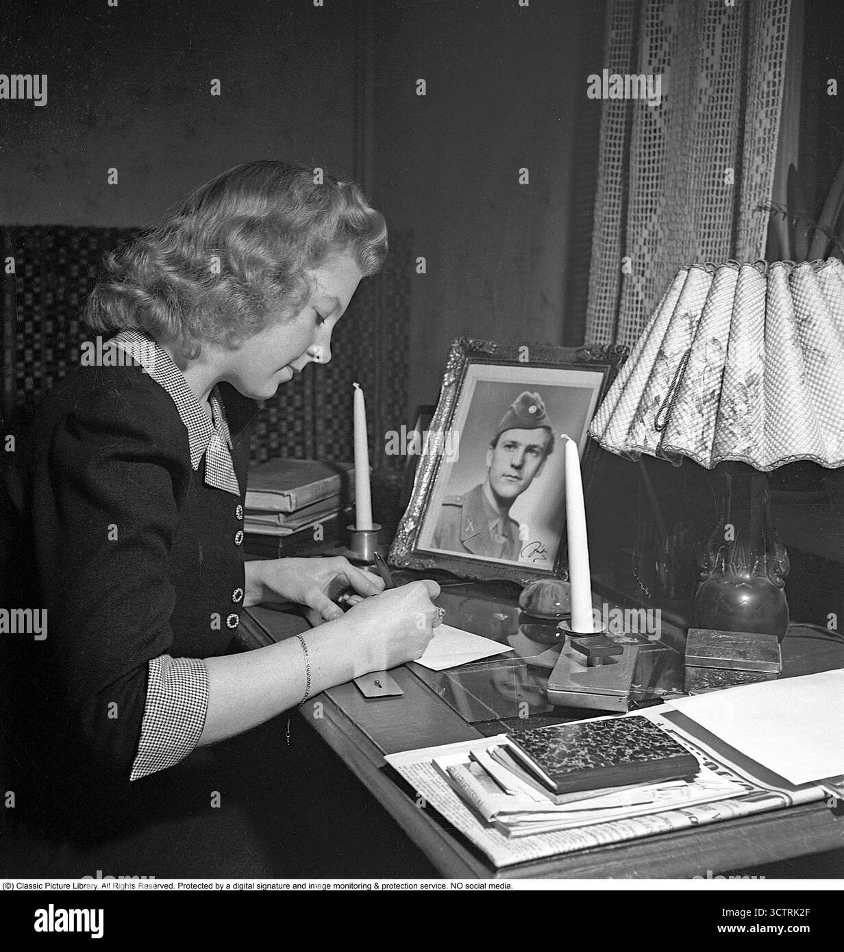 Nel 1945, una giovane donna si siede ad una scrivania ordinata, scrivendo una lettera. La busta è indirizzata al suo fidanzato, raffigurato in un'uniforme militare in una fotografia sulla scrivania, probabilmente arruolato per il servizio nell'ultimo anno della seconda guerra mondiale. Durante la seconda guerra mondiale, era comune per le donne scrivere lettere di cuore ai loro fidanzati che prestavano servizio in coscrizione militare. Queste regolari corrispondenze, piene di sentimenti personali e notizie da casa, mantenevano legami emotivi tra le incertezze della guerra, offrendo conforto e connessione su distanze. (Kristoffersson rif. R60-3) Foto Stock