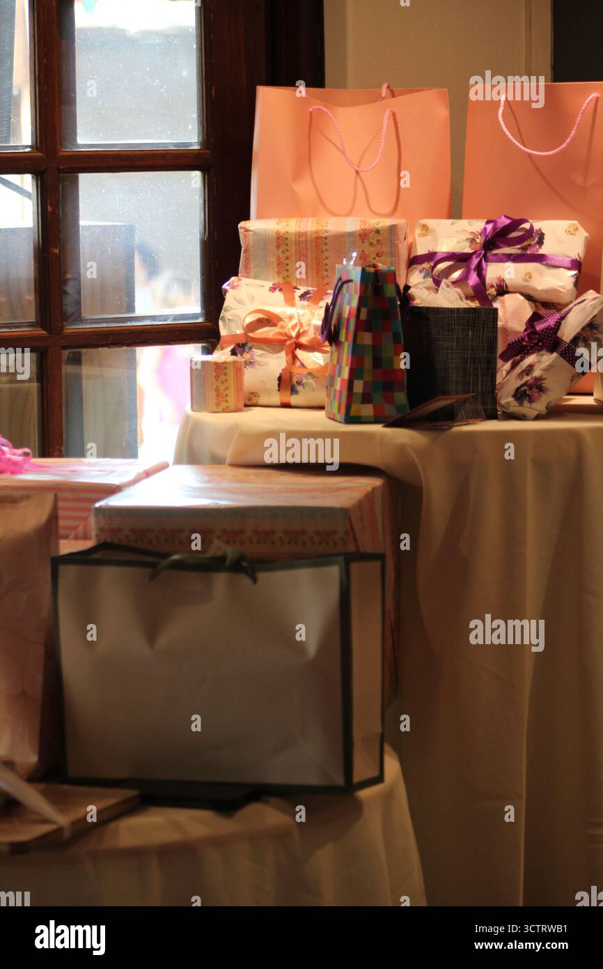Collezione di confezioni regalo confezionate e borse regalo colorate disposte su un tavolo vicino a una finestra, illuminate da una calda luce naturale durante una celebrazione o un evento Foto Stock