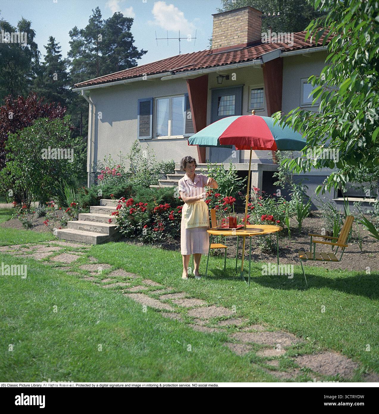 Nel 1958, una donna è ritratta nel giardino della sua casa Borohus, un simbolo del comfort domestico svedese. Borohus, un'azienda di costruzione di case che risale alla fine del XIX secolo, crebbe con il movimento della casa degli anni '1920. Fondata a Landsbro, Småland, il suo nome, abbreviazione di "Bo i ro i eget bo" (vivere pacificamente nella tua casa), rifletteva la sua etica. Come principale produttore svedese di case prefabbricate, Borohus prosperò con moduli, pareti e interni costruiti in fabbrica, tra cui cucine e armadi. Acquisita dalla cooperativa HSB nel 1936, ha operato fino al 1984. Nonostante la successiva bancarotta Foto Stock