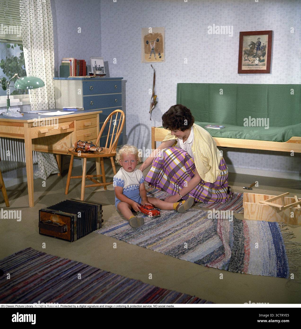 Nel 1958, una donna e suo figlio giocano gioiosamente con un'auto giocattolo sul pavimento di una casa Borohus, simbolo del comfort domestico svedese. Borohus, un'azienda di costruzione di case che risale alla fine del XIX secolo, crebbe con il movimento della casa degli anni '1920. Fondata a Landsbro, Småland, il suo nome, abbreviazione di "Bo i ro i eget bo" (vivere pacificamente nella tua casa), rifletteva la sua etica. Come principale produttore svedese di case prefabbricate, Borohus prosperò con moduli, pareti e interni costruiti in fabbrica, tra cui cucine e armadi. Acquisita dalla cooperativa HSB nel 1936, ha operato fino al 1984. Foto Stock