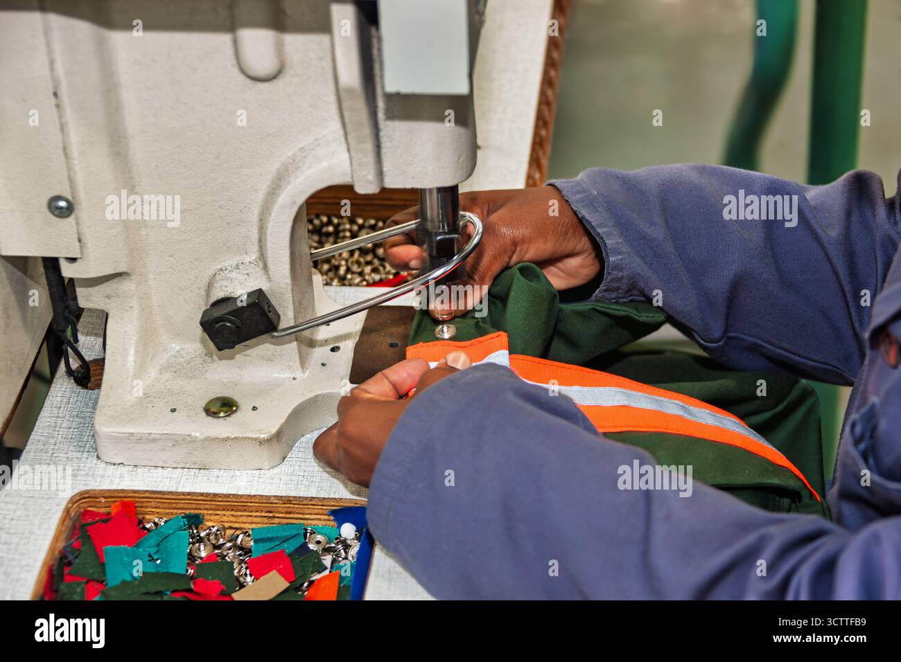 Eyeleting machine rivettatrice meccanica pesante operaio africano nella fabbrica tessile che produce indumenti protettivi da lavoro Foto Stock