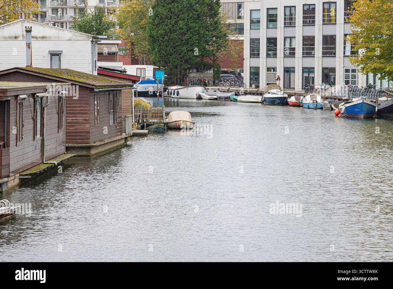 Vista sul canale di Amsterdam con casa sull'acqua e piccola barca sulla sinistra, grattacieli in costruzione e edifici residenziali tra gli alberi sulla piattaforma Foto Stock