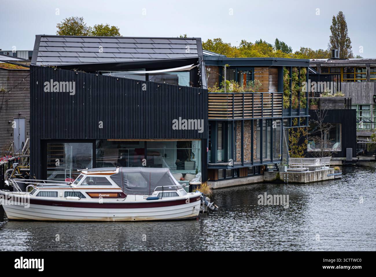 Vista sul canale di Amsterdam con casa sull'acqua e piccola barca sulla sinistra, grattacieli in costruzione e edifici residenziali tra gli alberi sulla piattaforma Foto Stock