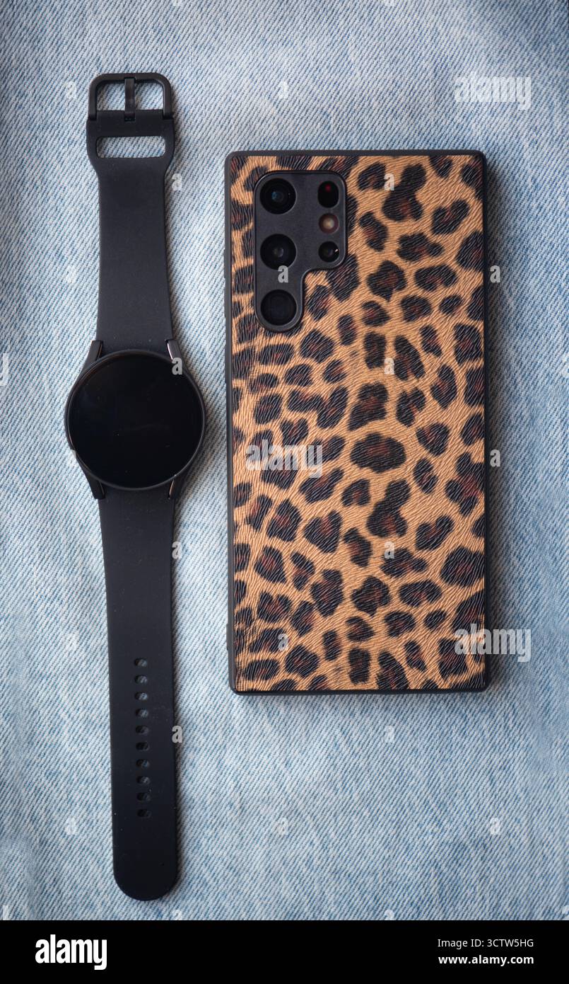 Custodia per smartphone con stampa leopardata e smartwatch nero su tessuto denim, che mettono in risalto gli accessori tecnologici alla moda in un ambiente casual Foto Stock
