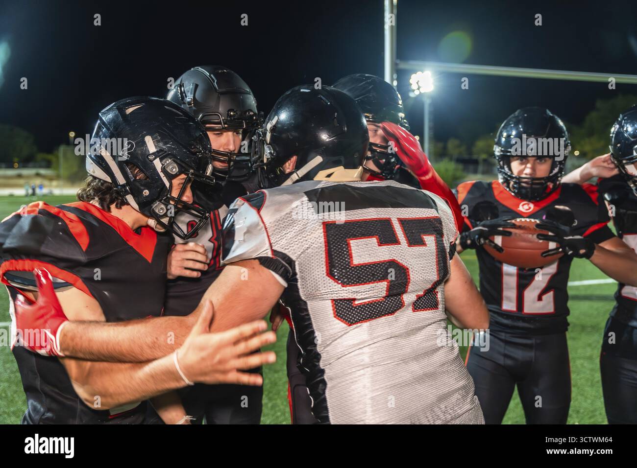 Giocatori di football americano in uniforme nera e rossa con caschi e spalline che si accoccolano su un campo di erba verde durante una partita notturna, discutendo di stra Foto Stock