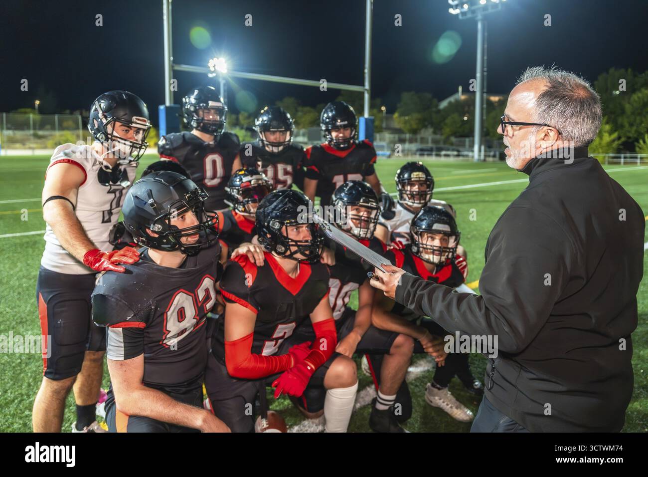 Allenatore di football americano che discute di strategia con la sua squadra di giocatori in uniforme e caschi durante una pratica notturna o una partita su un campo di erba verde Foto Stock
