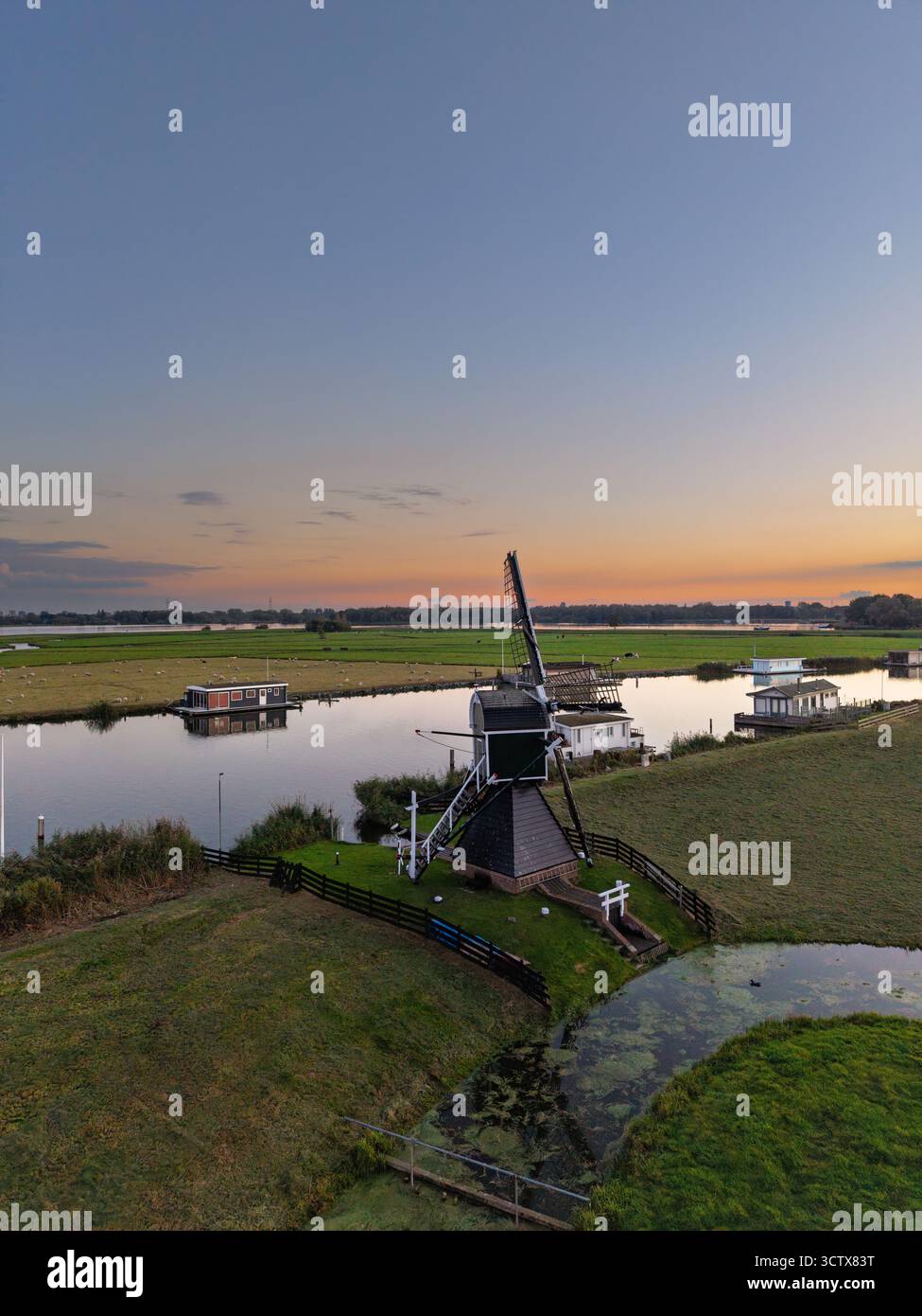 Vista aerea della campagna olandese mulino a vento verde scuro con accenti bianchi vicino al canale calmo, case galleggianti e recinzioni fiancheggiano le rive erbose, il cielo si riflette al tramonto Foto Stock