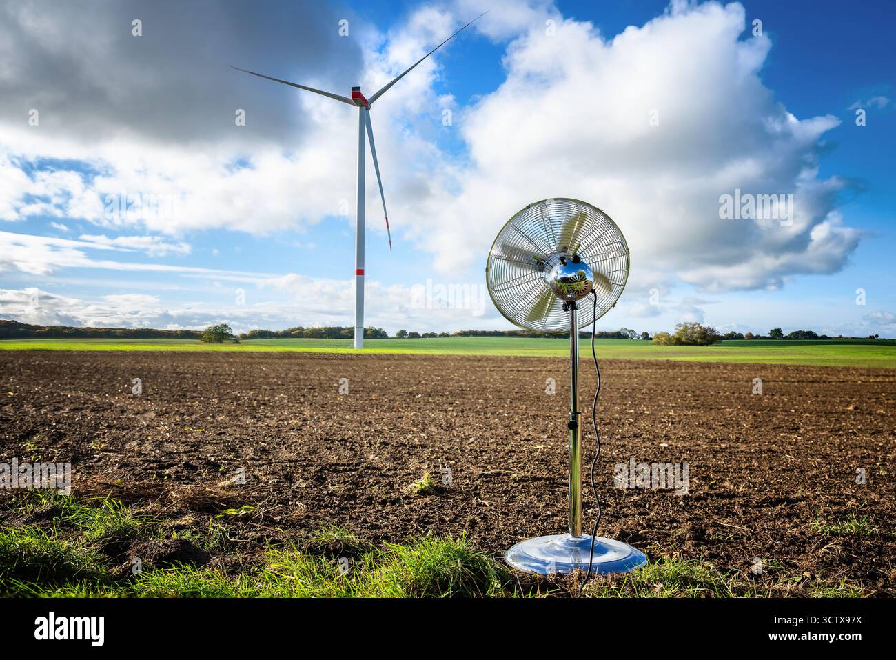 Ventilatore domestico d'argento in piedi su un ampio campo di fronte a un'alta turbina eolica, concetto di progresso e tecnologia, cielo blu con nuvole, copia spazio Foto Stock