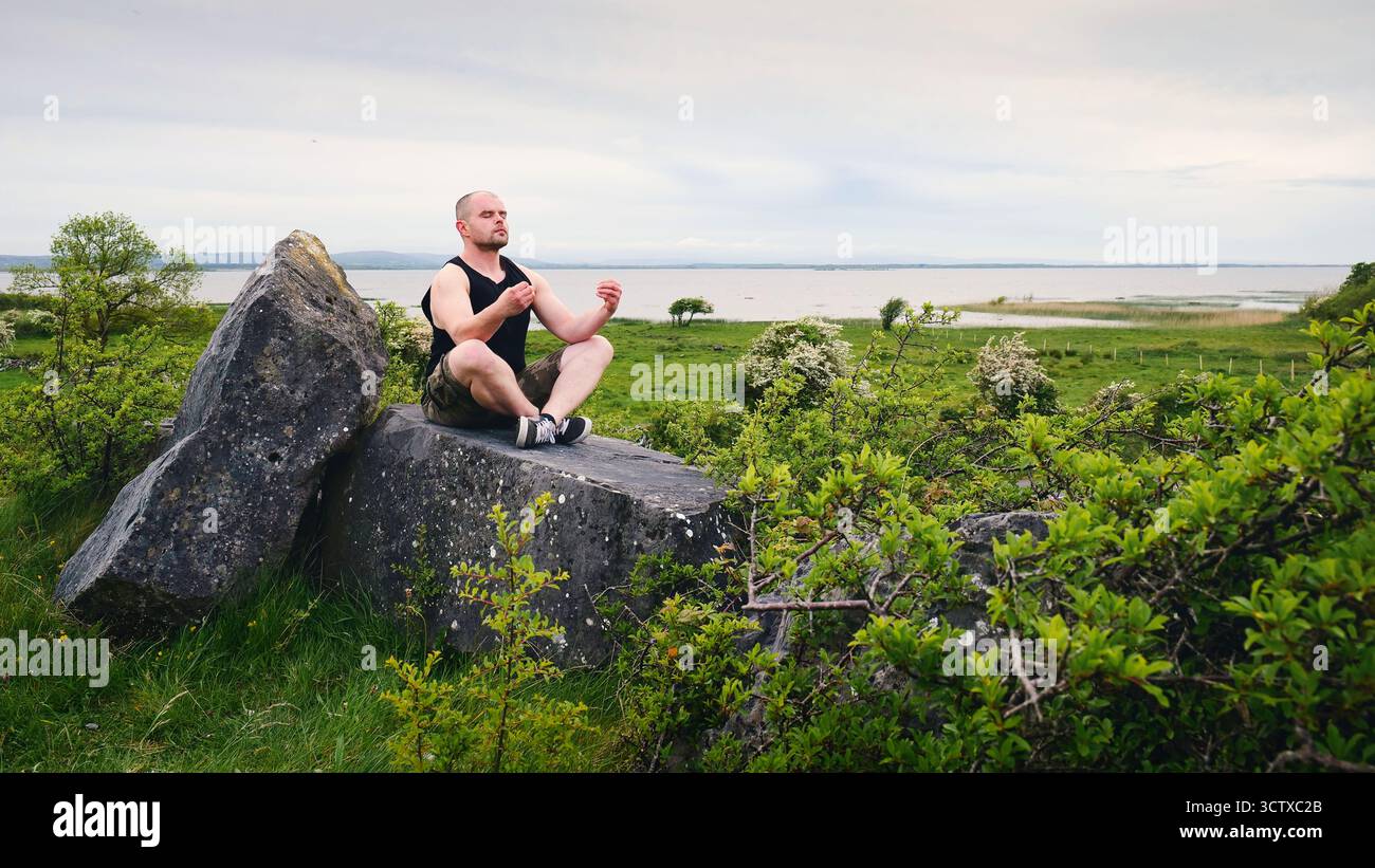 Uomo meditando all'aperto su rocce vicino a Galway costa Irlanda, praticare consapevolezza, yoga, relax, salute mentale, benessere, libertà e natura Foto Stock