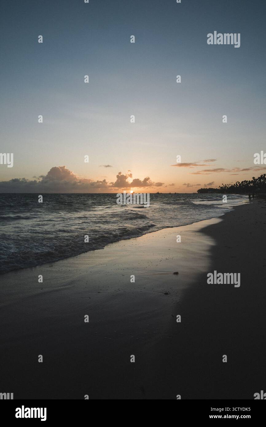 Onde dolci si infrangono sulla riva mentre il sole tramonta all'orizzonte, generando colori caldi sul cielo. La tranquilla spiaggia invita a riflettere tranquillamente al crepuscolo Foto Stock
