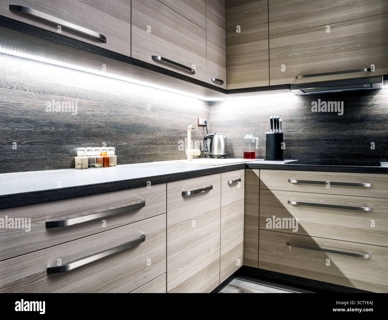 Cucina moderna e luminosa con armadi in legno chiaro e piano di lavoro e spruzzi scuri, calde luci sotto il mobile che mettono in risalto lavandino e fornelli per un ambiente accogliente Foto Stock