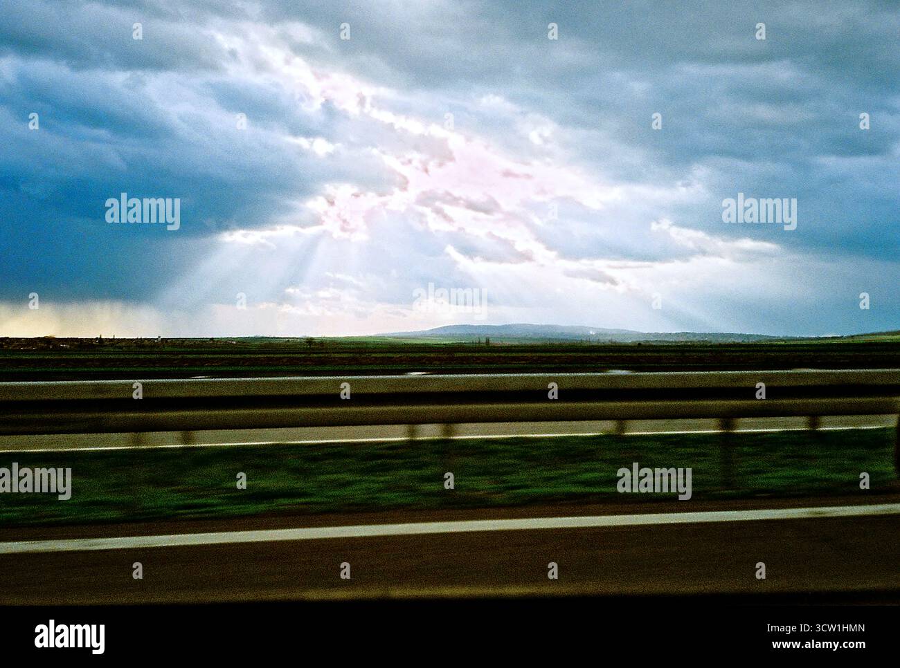 Foto analogica da 35 mm dal lato del conducente che mostra la recinzione autostradale e il cielo coperto con raggi di luce che irrompono. Pomeriggio nuvoloso, atmosfera d'archivio. Foto Stock