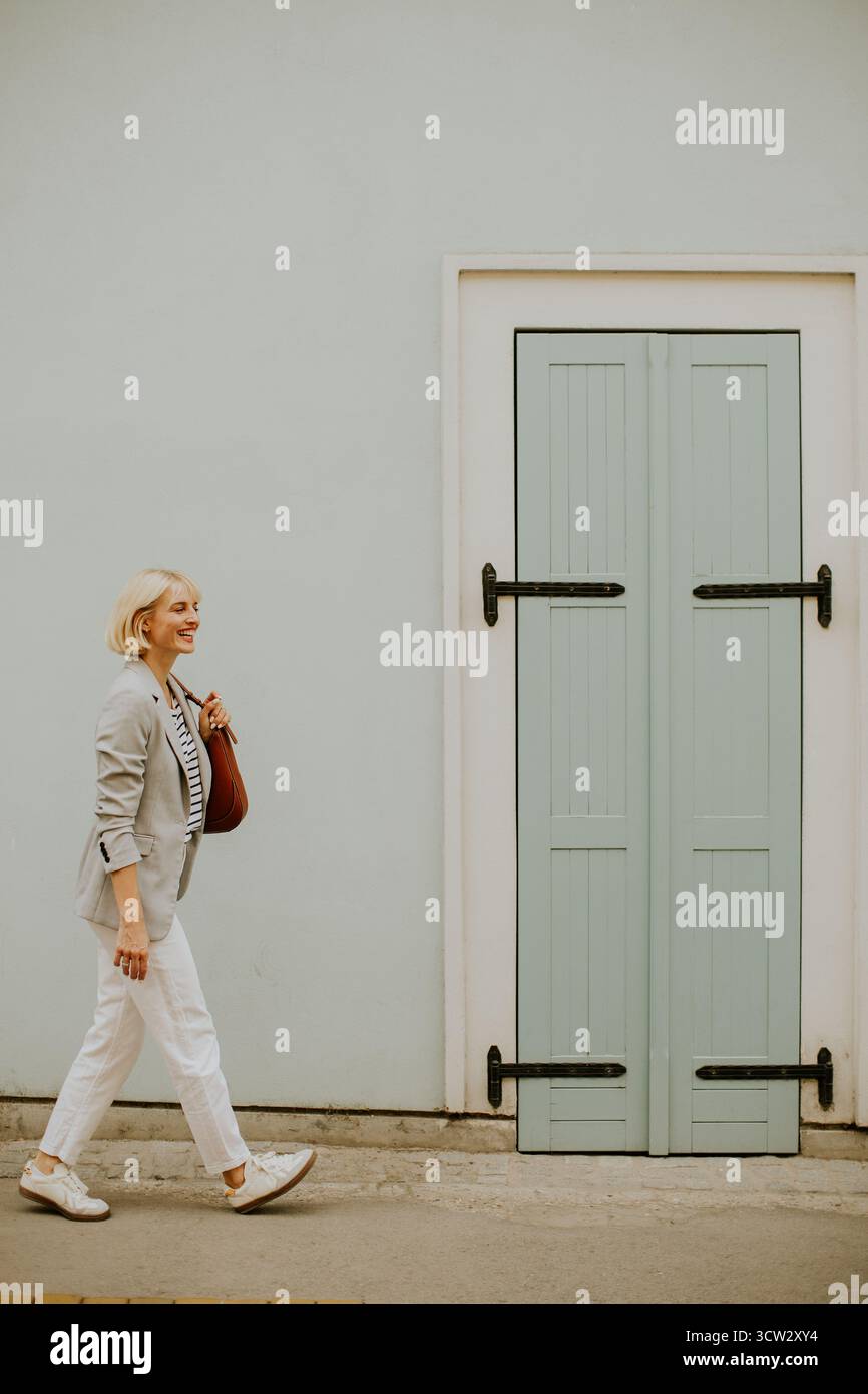 Una donna cammina in modo informale lungo la strada, indossando un outfit rilassato con pantaloni bianchi e un blazer grigio. Porta con sé una borsa marrone mentre si gode la sua giornata Foto Stock