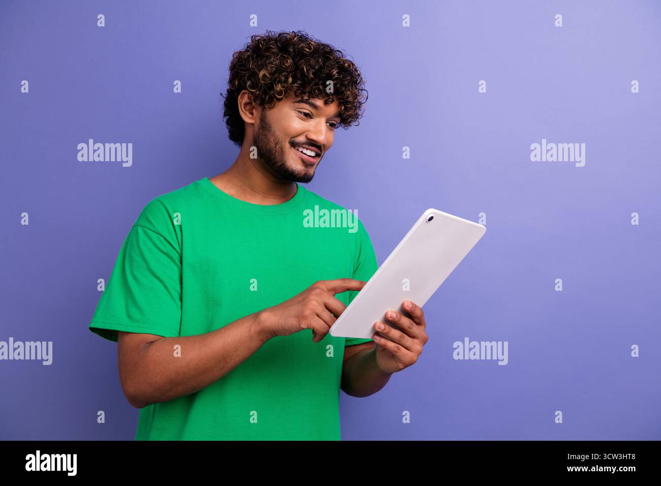 Il giovane con i capelli ricci usa un tablet sorridente su uno sfondo viola e indossa una t-shirt verde, sottolineando la tecnologia e il tempo libero Foto Stock