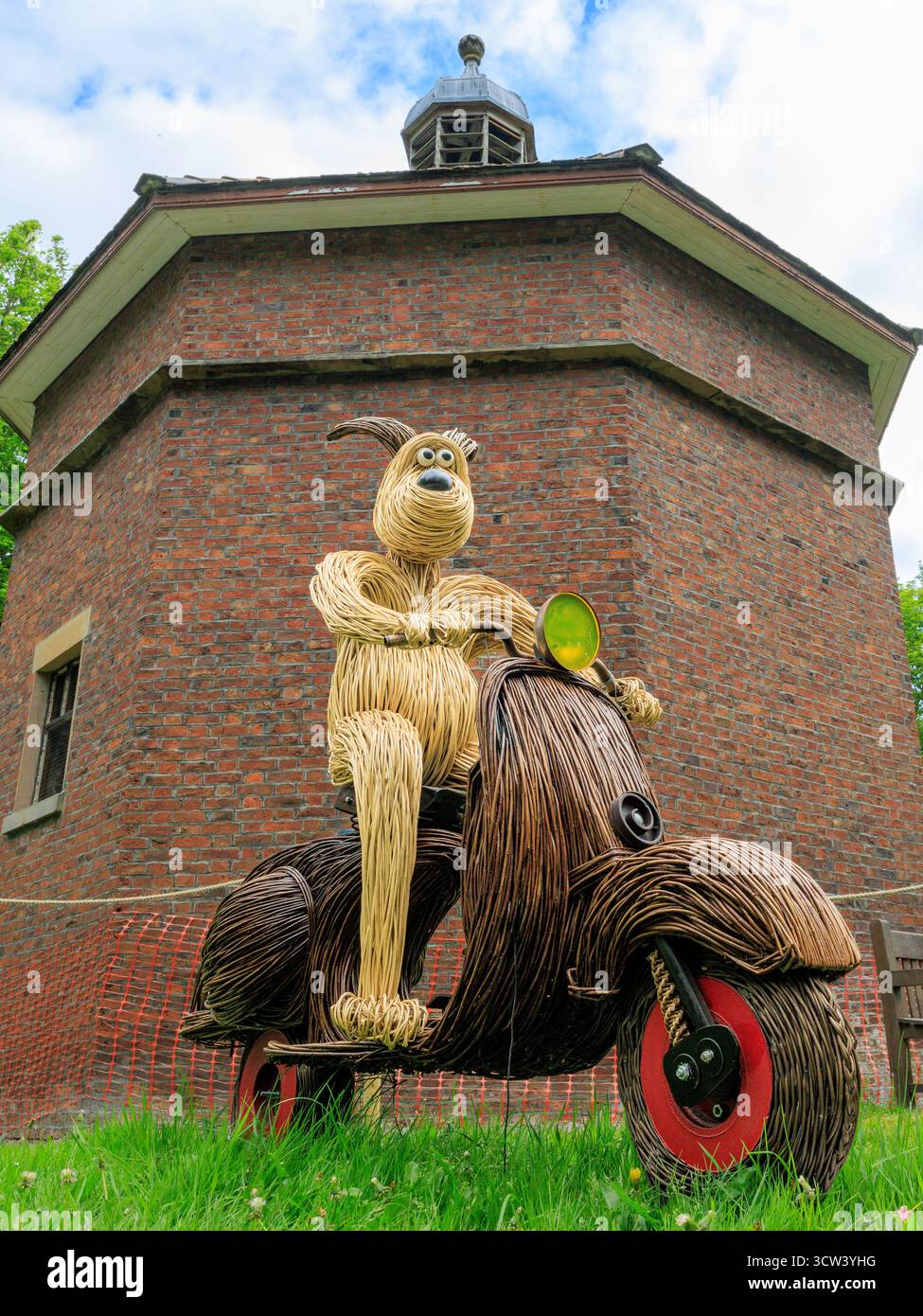 scultura in vimini realizzata a mano di un cane da passeggio in sella a uno scooter di fronte alla storica colomba ottagonale in mattoni nei terreni di lytham hall Foto Stock