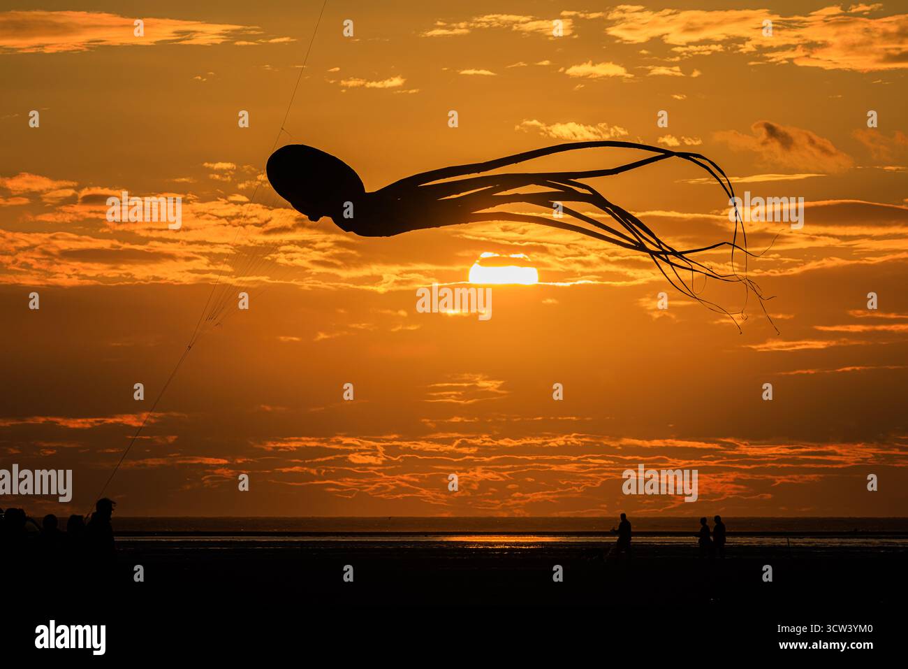 una suggestiva silhouette di un grande aquilone di polpo gonfiabile con lunghi tentacoli trainanti in un vivace cielo nuvoloso al tramonto arancione che si riflette in un mare calmo Foto Stock