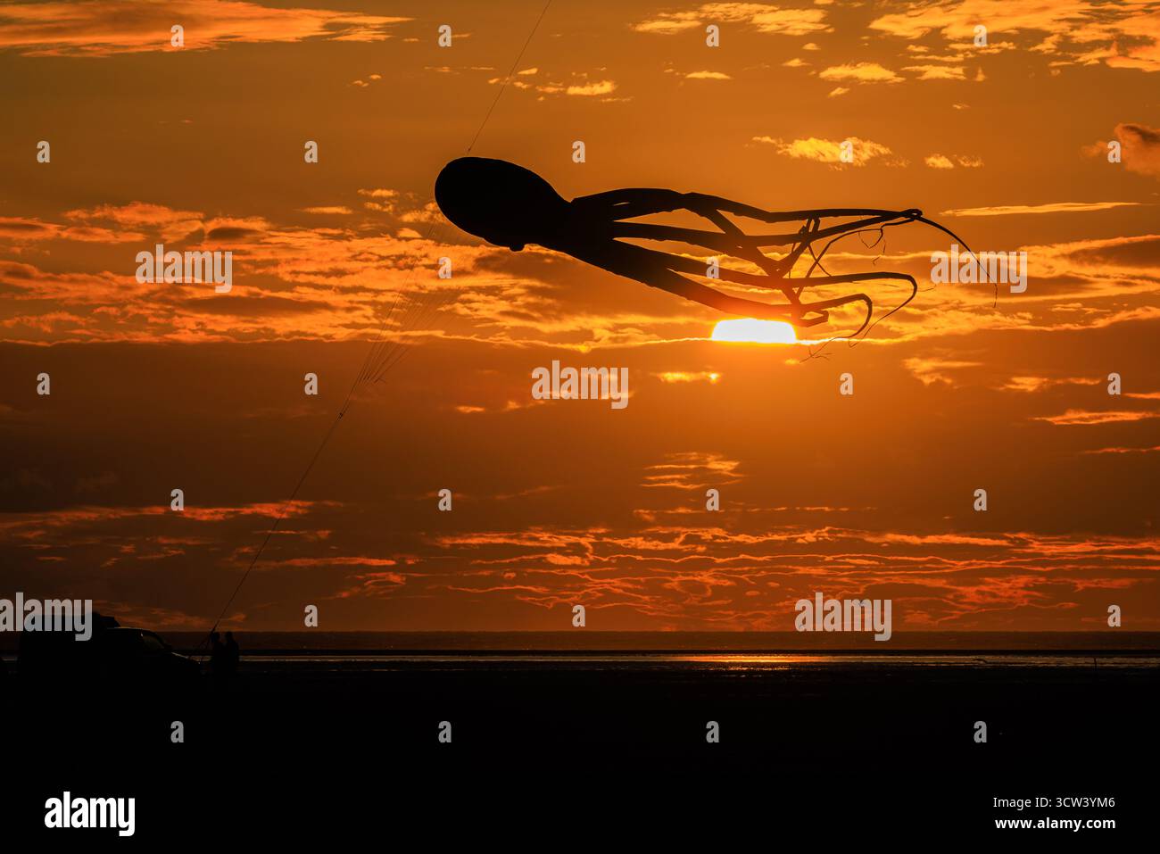 una suggestiva silhouette di un grande aquilone di polpo gonfiabile con lunghi tentacoli trainanti in un vivace cielo nuvoloso al tramonto arancione che si riflette in un mare calmo Foto Stock