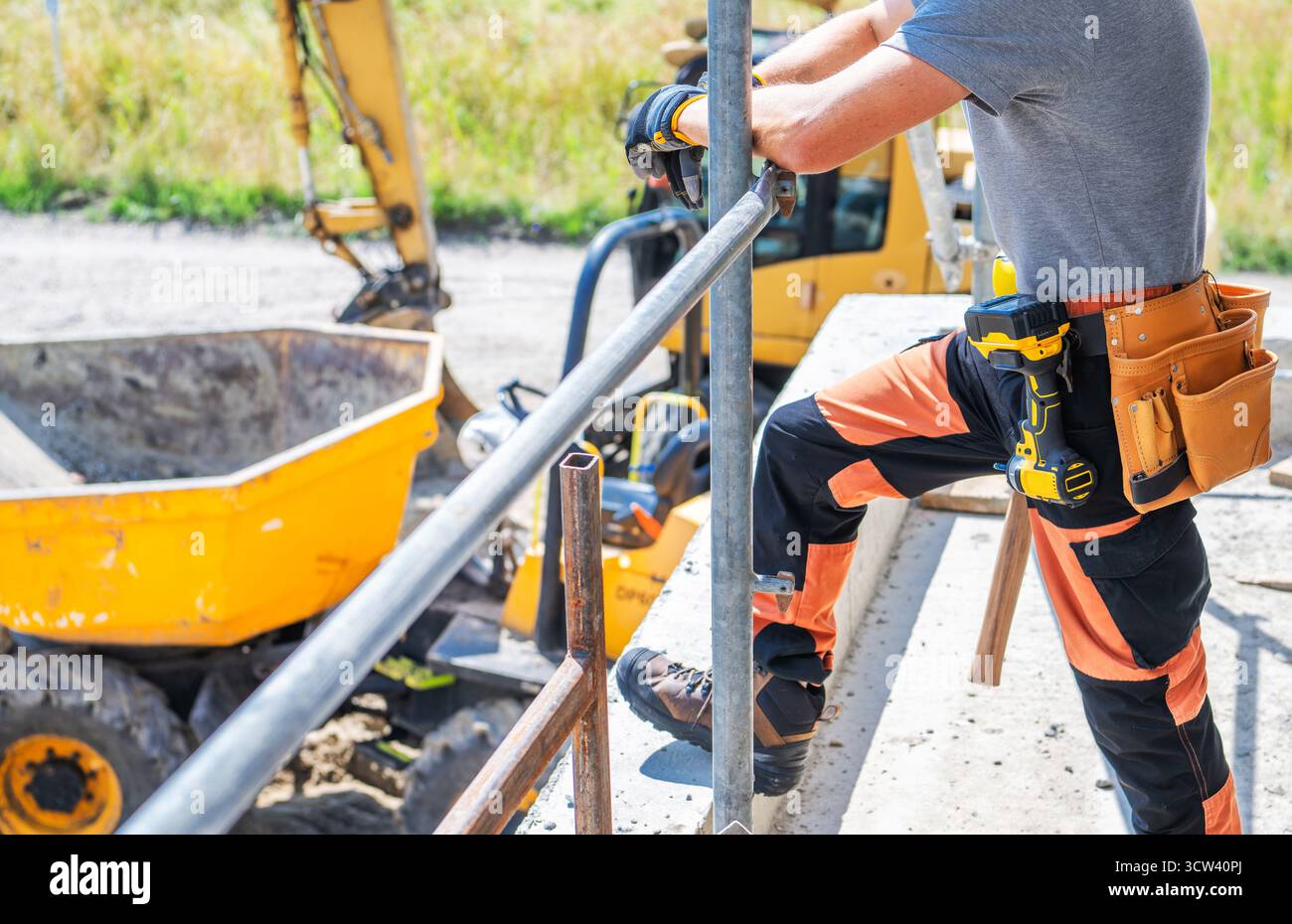 Un operaio edile si trova su una struttura, utilizzando utensili elettrici per svolgere attività mentre i macchinari gialli funzionano in background sotto il cielo limpido Foto Stock