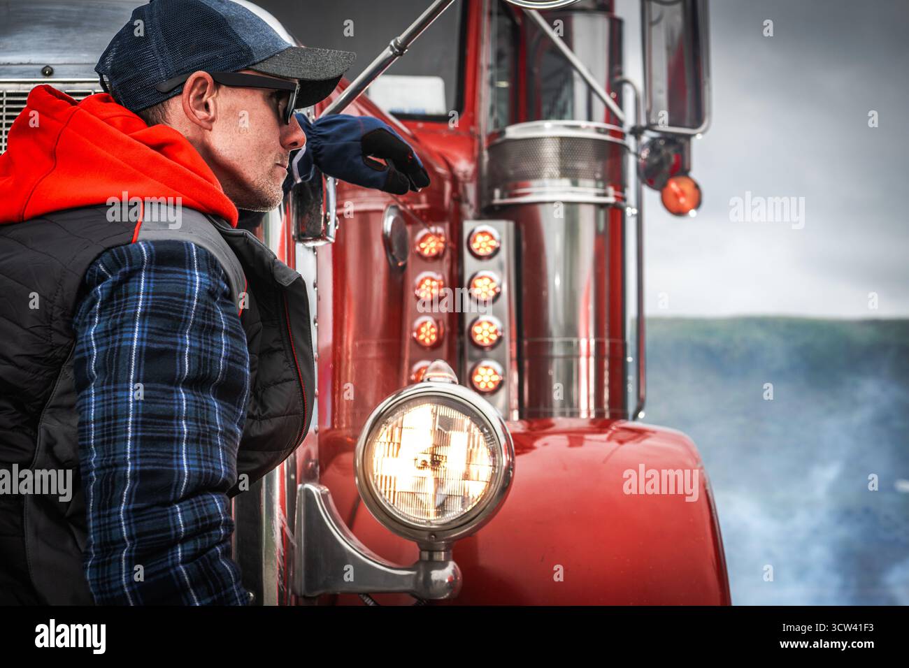 Un camionista con una camicia a scacchi blu e una felpa con cappuccio rossa si appoggia a un classico camion rosso, incentrato sull'orizzonte. Il clima fresco contribuisce all'atmosfera Foto Stock