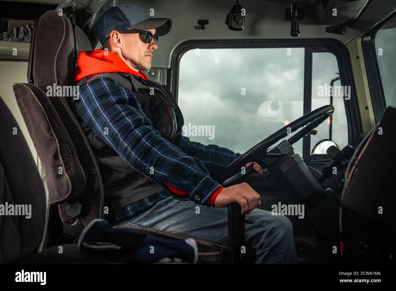 Un conducente esperto siede in sicurezza nella cabina di un camion pesante, indossando occhiali da sole e un berretto, mentre naviga attraverso un ambiente nuvoloso. Foto Stock