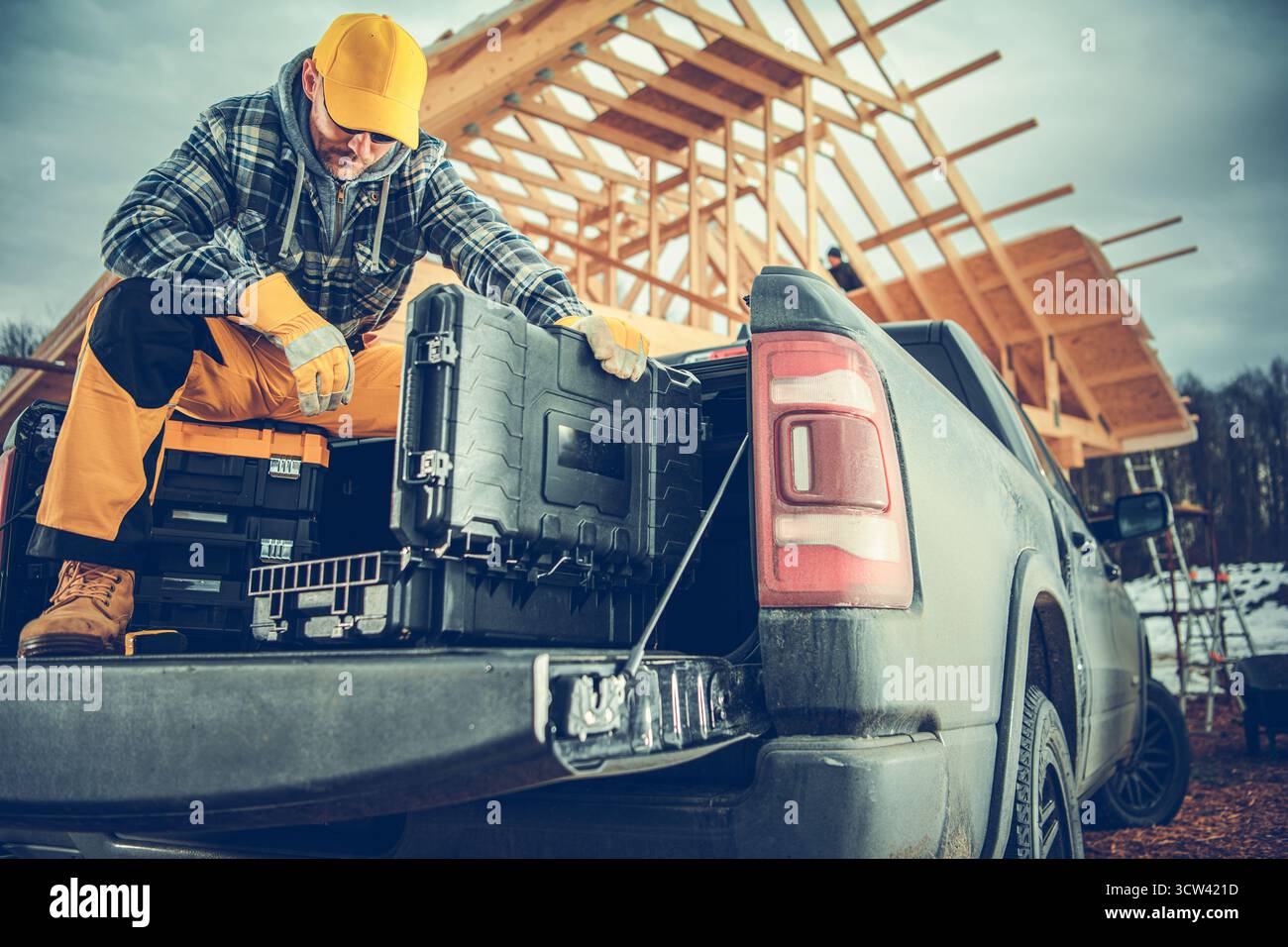 Un operaio edile con abiti caldi dispone gli attrezzi nel pianale del camion mentre lavora in cantiere. La neve può essere vista sullo sfondo. Foto Stock