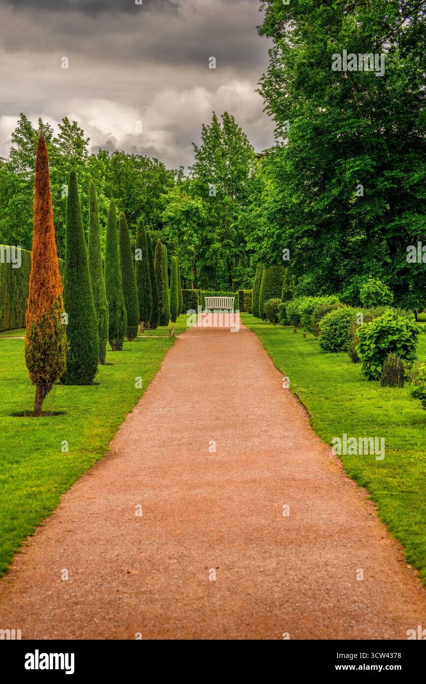 Bellissimo percorso del giardino inglese con vegetazione lussureggiante, alberi curati e una panca bianca sotto un cielo nuvoloso Foto Stock