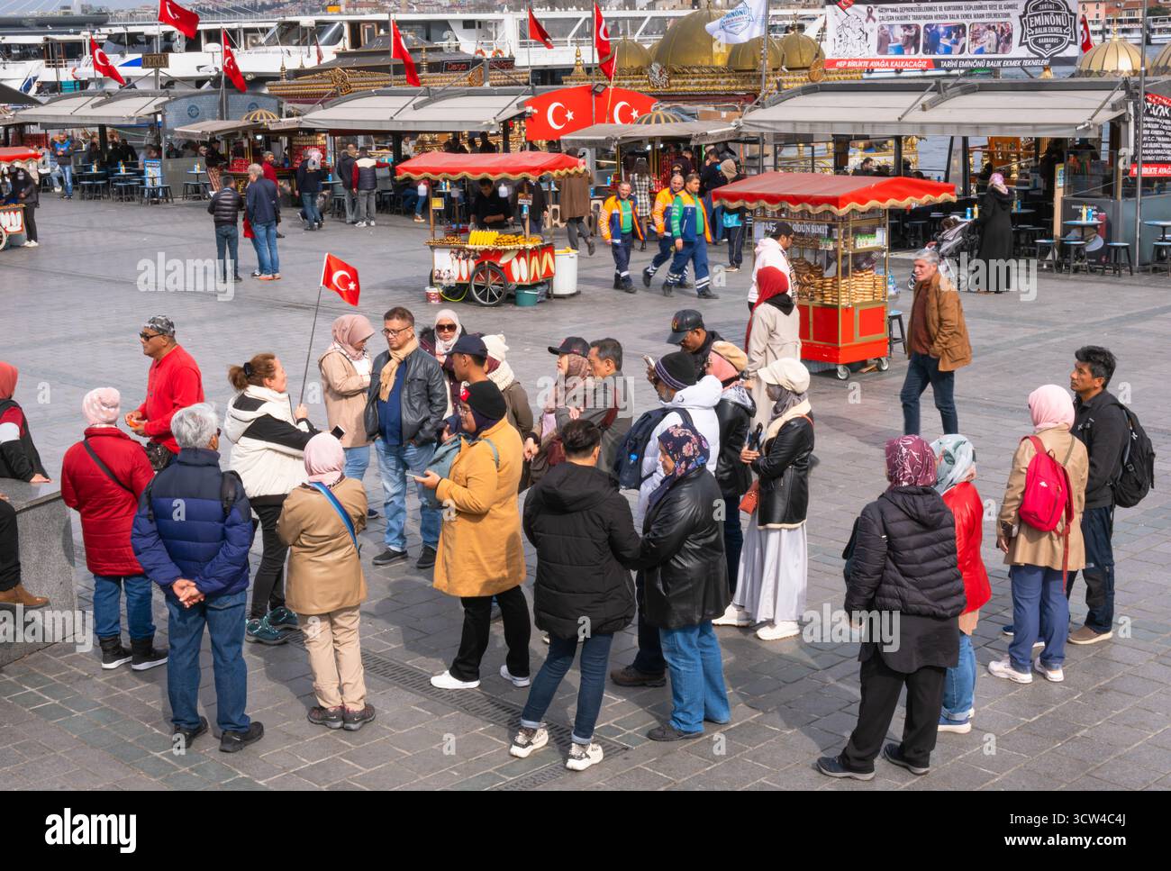 Istanbul, Turchia - 10 marzo 2023: Un gruppo di turisti si riunisce intorno alla guida del tour in Piazza Eminonu. La guida con bandiera turca spiega la storia Foto Stock