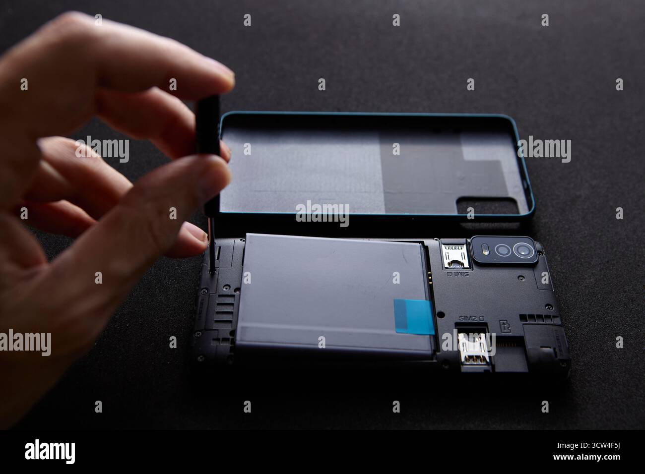 Primo piano di una mano che tiene un cacciavite sopra uno smartphone smontato su una superficie nera. La scena mostra l'inizio di un processo di riparazione con p Foto Stock