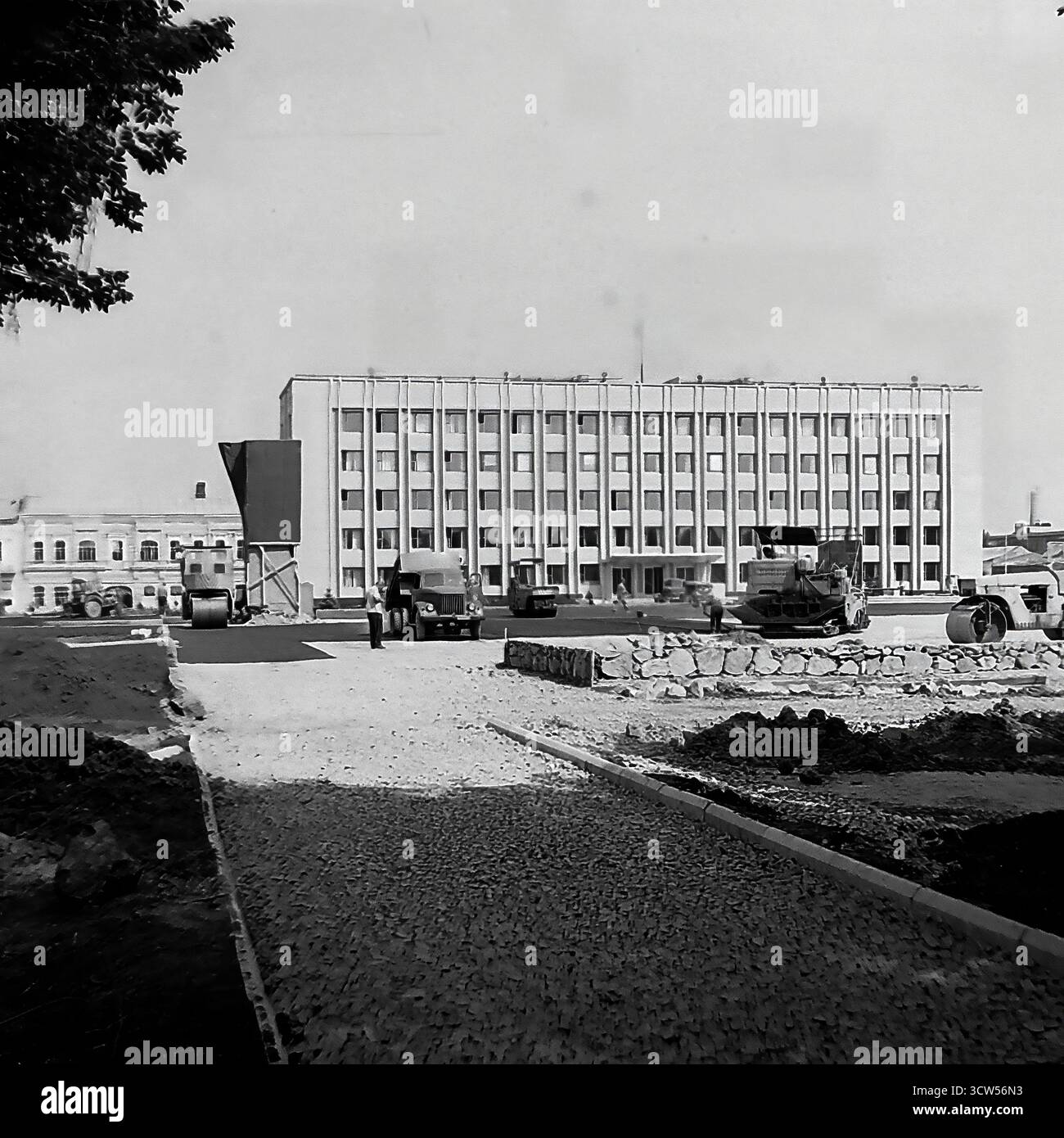 Un massiccio progetto di ricostruzione è in corso presso Piazza della Rivoluzione d'ottobre (ora Piazza Soborna), la piazza principale della città di Sloviansk, RSS Ucraina, in questa foto del 1980. La vasta area è un cantiere molto trafficato, con lavoratori e macchinari che posano nuove lastre di pavimentazione. Sullo sfondo sono visibili il monumento centrale a Lenin e l'edificio principale dell'amministrazione cittadina. Questa immagine in bianco e nero documenta un momento significativo di rinnovamento urbano e trasformazione dello spazio pubblico in una tipica città sovietica Foto Stock