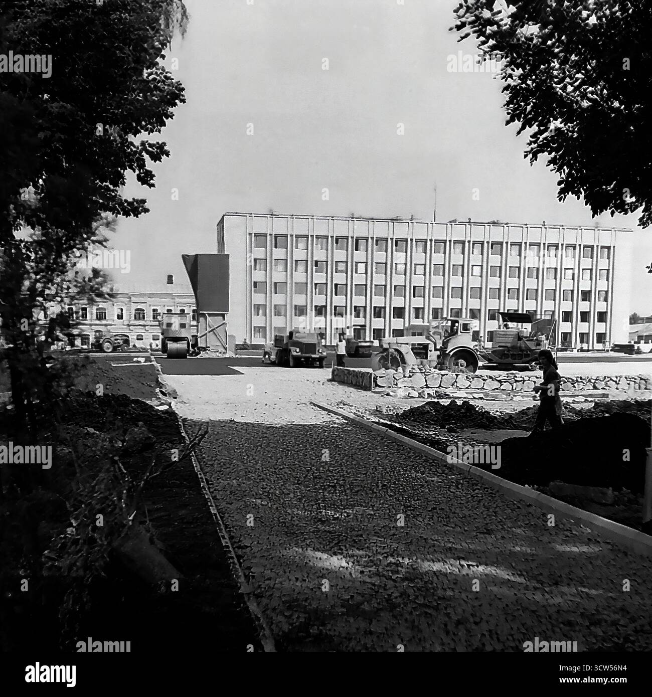 Un massiccio progetto di ricostruzione è in corso presso Piazza della Rivoluzione d'ottobre (ora Piazza Soborna), la piazza principale della città di Sloviansk, RSS Ucraina, in questa foto del 1980. La vasta area è un cantiere molto trafficato, con lavoratori e macchinari che posano nuove lastre di pavimentazione. Sullo sfondo sono visibili il monumento centrale a Lenin e l'edificio principale dell'amministrazione cittadina. Questa immagine in bianco e nero documenta un momento significativo di rinnovamento urbano e trasformazione dello spazio pubblico in una tipica città sovietica Foto Stock