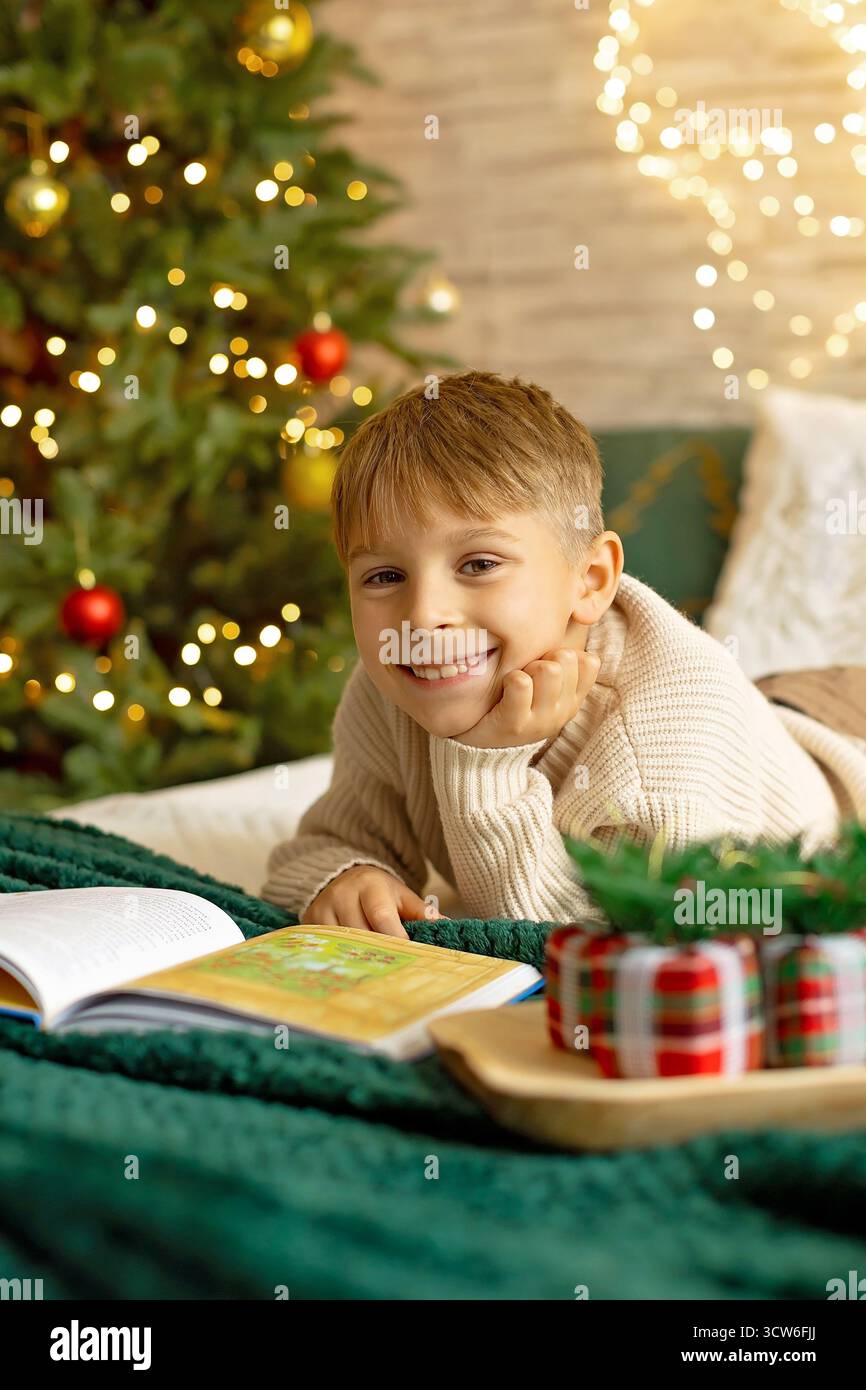 Adorabile bambino in età prescolare, seduto davanti al caminetto, tenendo in mano il regalo di natale e leggendo un libro, gioia e felicità a casa Foto Stock