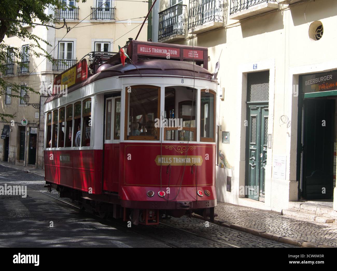Tram rosso su strada acciottolata a Lisbona, Portogallo - Un tram turistico rosso d'epoca (Hills Tramcar Tour) che si sposta lungo una strada acciottolata incorniciata dalla luce Foto Stock