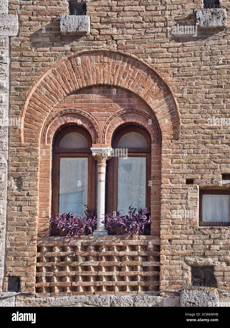 Finestra medievale in mattoni con piantatrice - primo piano di una finestra rustica in mattoni ad arco con colonna centrale in pietra, caratterizzata da una scatola di fioriera di colore viola Foto Stock
