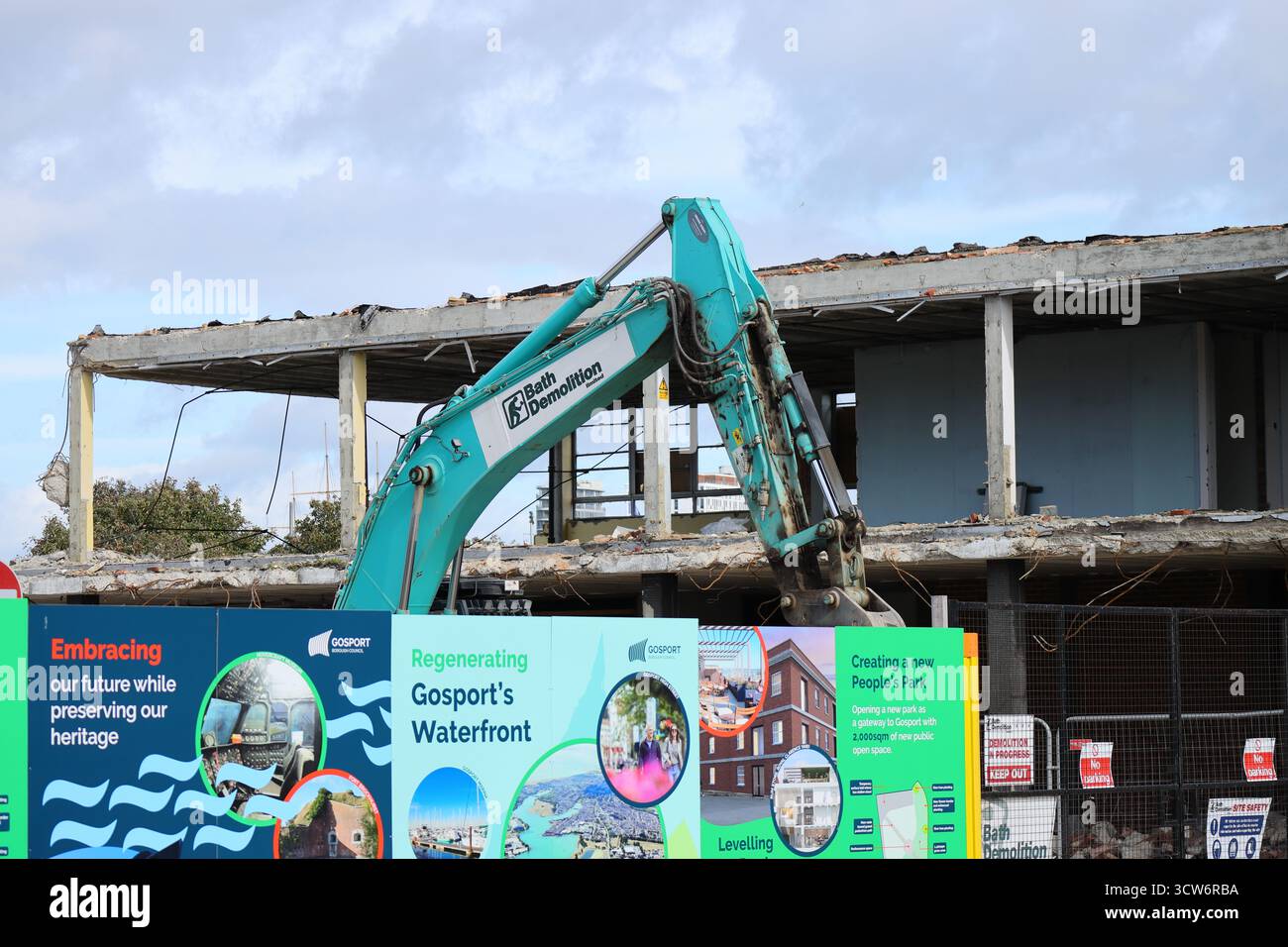 Demolizione in corso dell'ex stazione degli autobus di Gosport, Hampshire, Inghilterra. Un escavatore verde si erge davanti a una struttura scheletrica in fase di demolizione, incorniciata da un vibrante cartello che promuove la rigenerazione del lungomare di Gosport. Il tabellone presenta rendering architettonici e frasi come "creare un nuovo Parco delle persone", che combinano ambizione civica con la conservazione del patrimonio. Il contrasto tra il cemento esposto e la grafica visionaria cattura un momento di trasformazione urbana. Foto Stock