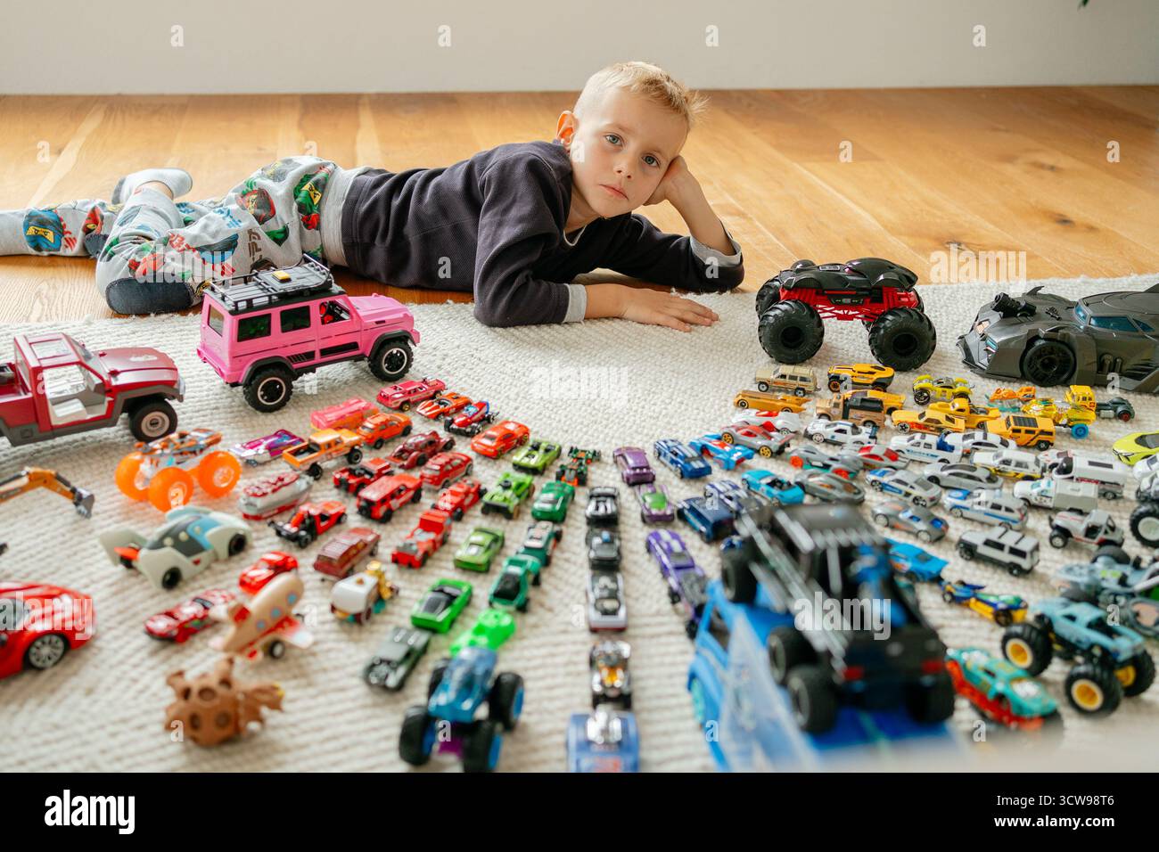 Vista dall'alto verso il basso di un ragazzo sdraiato sul pavimento, circondato da una vasta e colorata collezione di macchinine giocattolo disposte ordinatamente in file per hobby e giochi d'infanzia Foto Stock